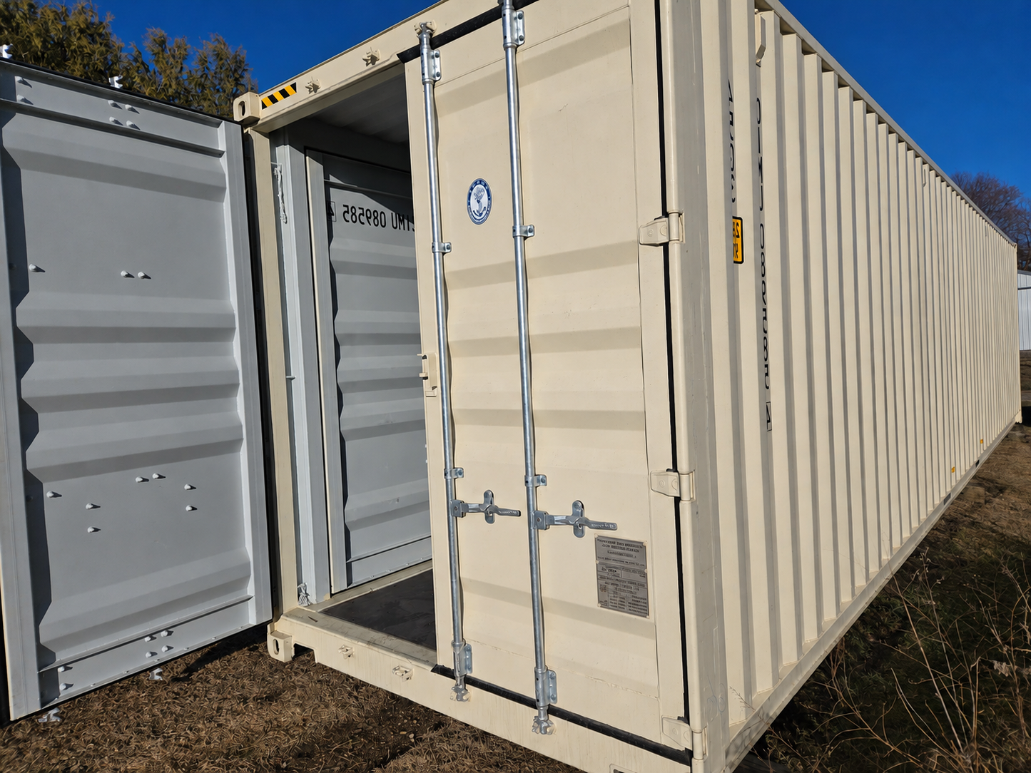 A large beige shipping container with its front door open, showing the interior and a gray outer door, photographed outdoors on grass with blue sky and trees in the background.